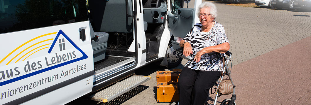 Eine Frau sitzt vor dem Kleinbus des Seniorenzentrums auf ihren Rollator.
