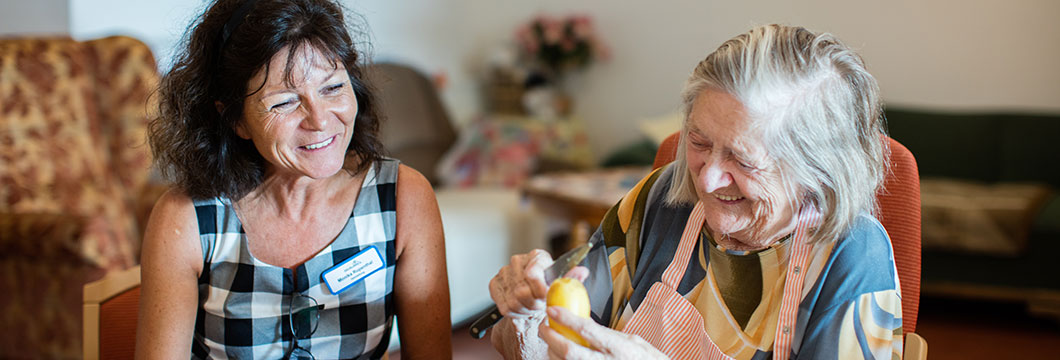 Eine Seniorin schält in fröhlicher Gesellschaft mit einer Pflegerin Kartoffeln.