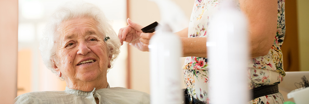 "Eine ältere Dame sitzt glücklich und frisch frisiert im Friseursalon des Seniorenzentrums."