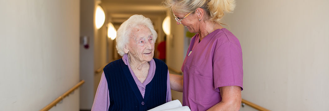 Im Gang des Seniorenzentrums legt eine Pflegerin freundlich die Hand auf den Rücken einer Seniorin.