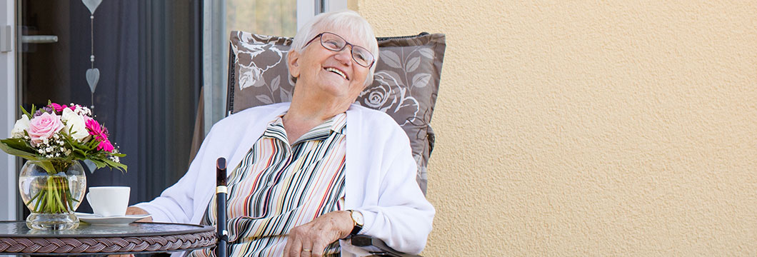 Eine Seniorin sitzt in einem bequemen Gartenstuhl auf einer Veranda vor dem Seniorenzentrum.