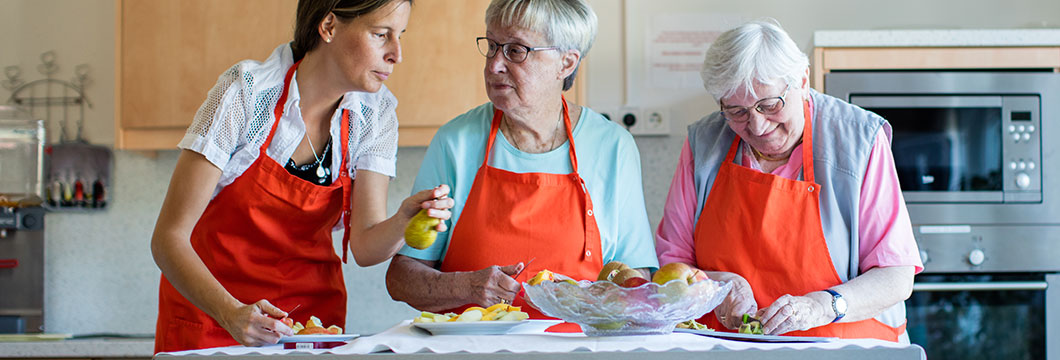 Zwei Seniorinnen schneiden gemeinsam mit einer Pflegerin Obst für einen Salat.