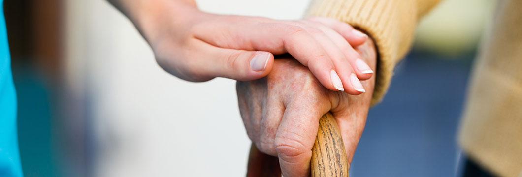 Die Hand einer Pflegerin liegt fürsorglich auf der eines Senioren, dessen Hand ein Gehstock umfasst.