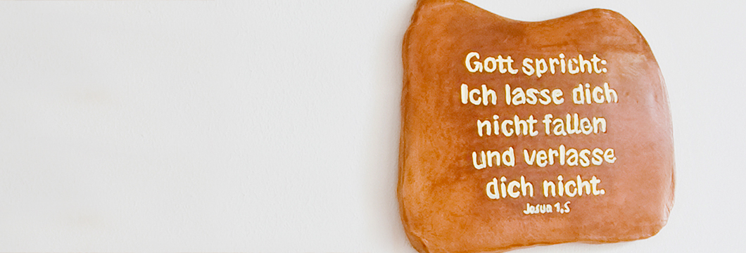 An einer weißen Wand hängt eine Keramiktafel mit dem Vers: Gott spricht: Ich lasse dich nicht fallen und verlasse dich nicht. Josua 1,5